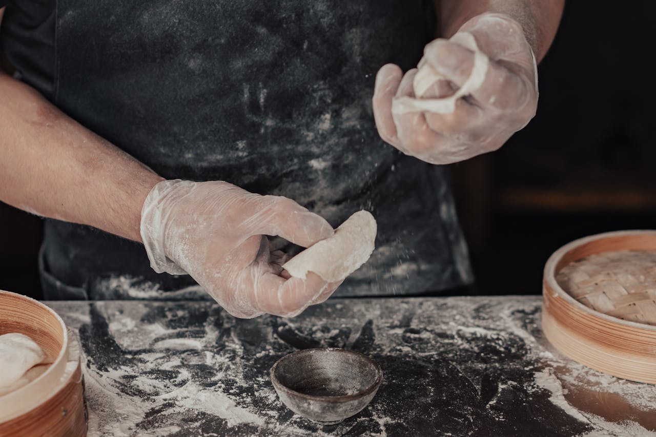 Home Close-up of a person making dumplings with a bamboo steamer, featuring flour and dough.